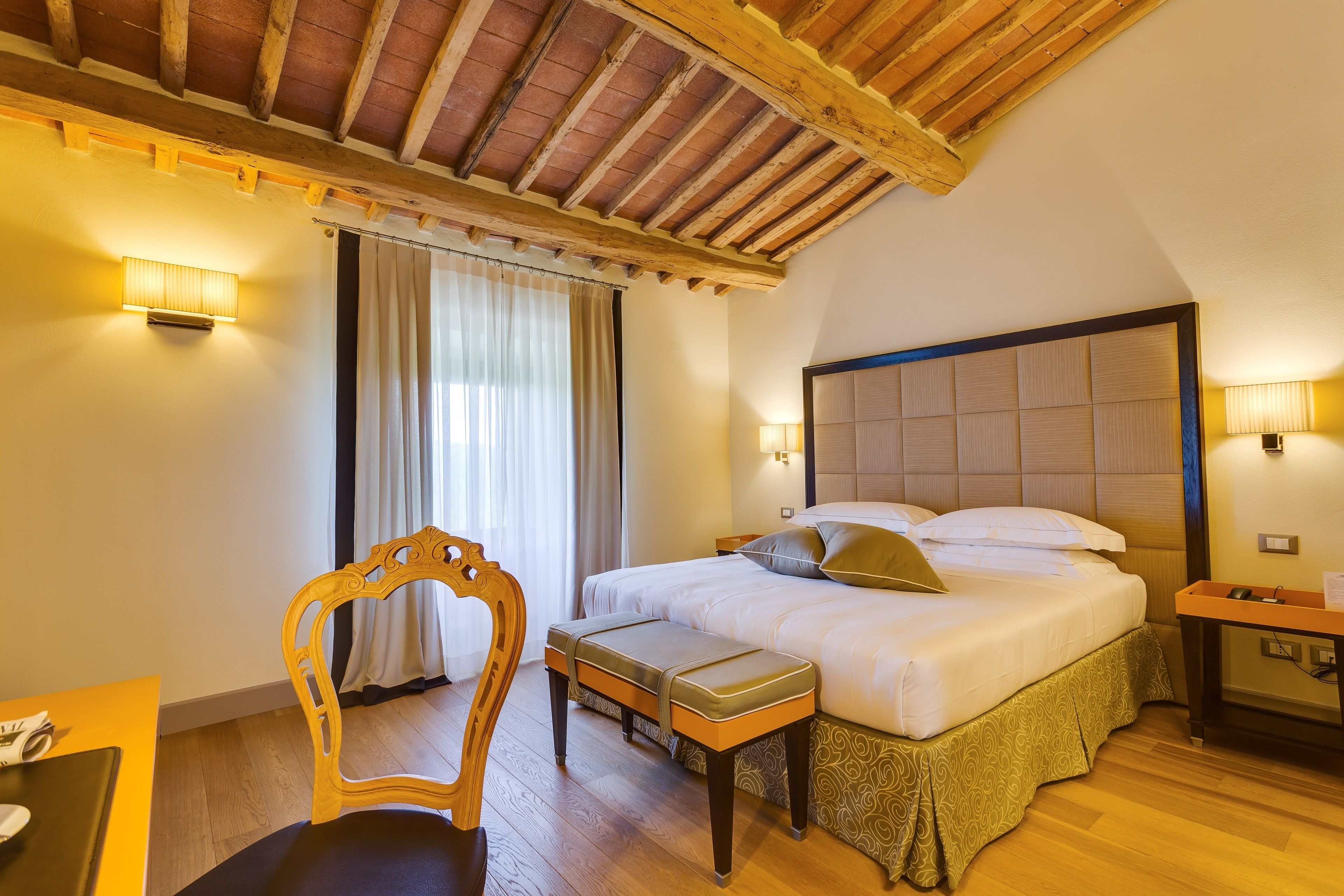 A hotel room with a wooden ceiling, a bed with a patterned bed skirt, a bedside table with a lamp, and a chair with a cushioned seat.