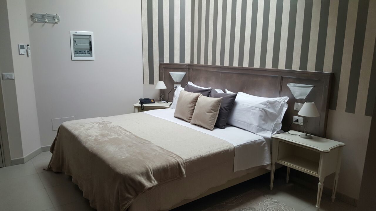 Una cama bien hecha con cubrecama y almohadas color beige, cabecera de madera, mesitas de noche con lámparas y un estampado a rayas en la pared.