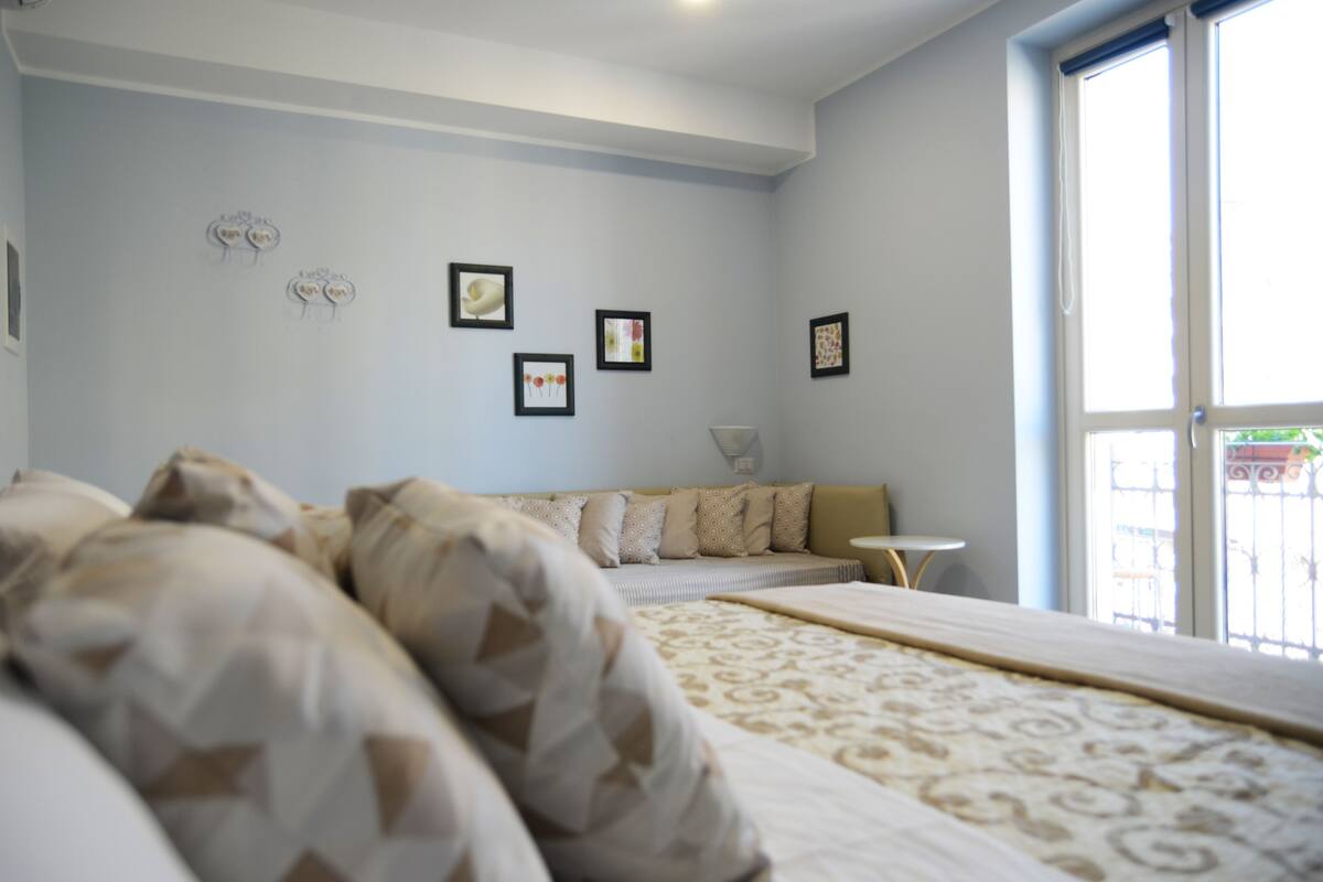 A hotel room with a large bed, a sofa, and a small table. There are framed pictures on the wall and a window with a view of the outside.