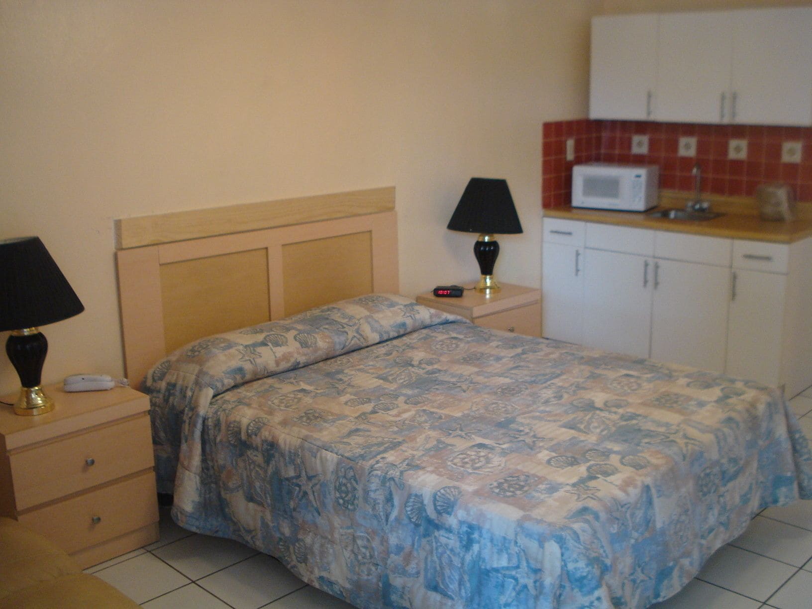 A bedroom with a bed, bedside tables, a lamp, a clock, and a kitchenette with a microwave.