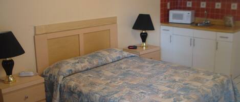 A bedroom with a bed, bedside tables, a lamp, a clock, and a kitchenette with a microwave.