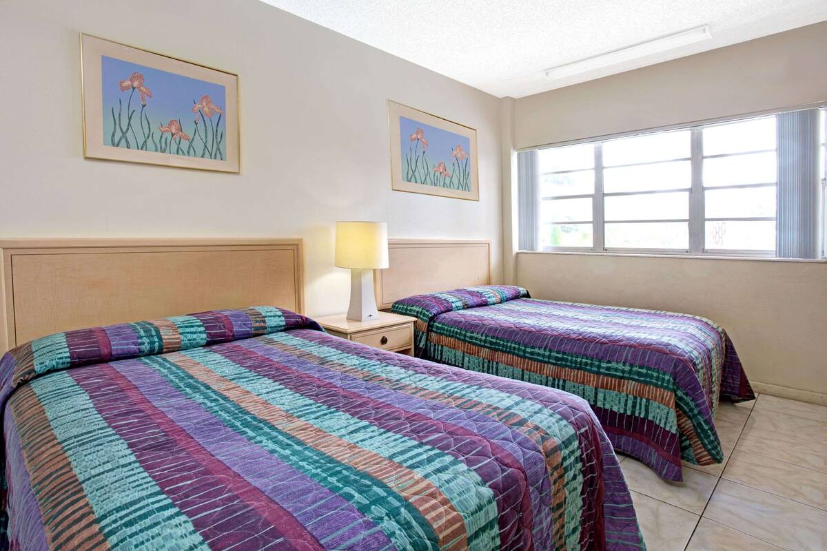 A hotel room with two beds, a nightstand with a lamp, and two framed floral paintings on the wall.