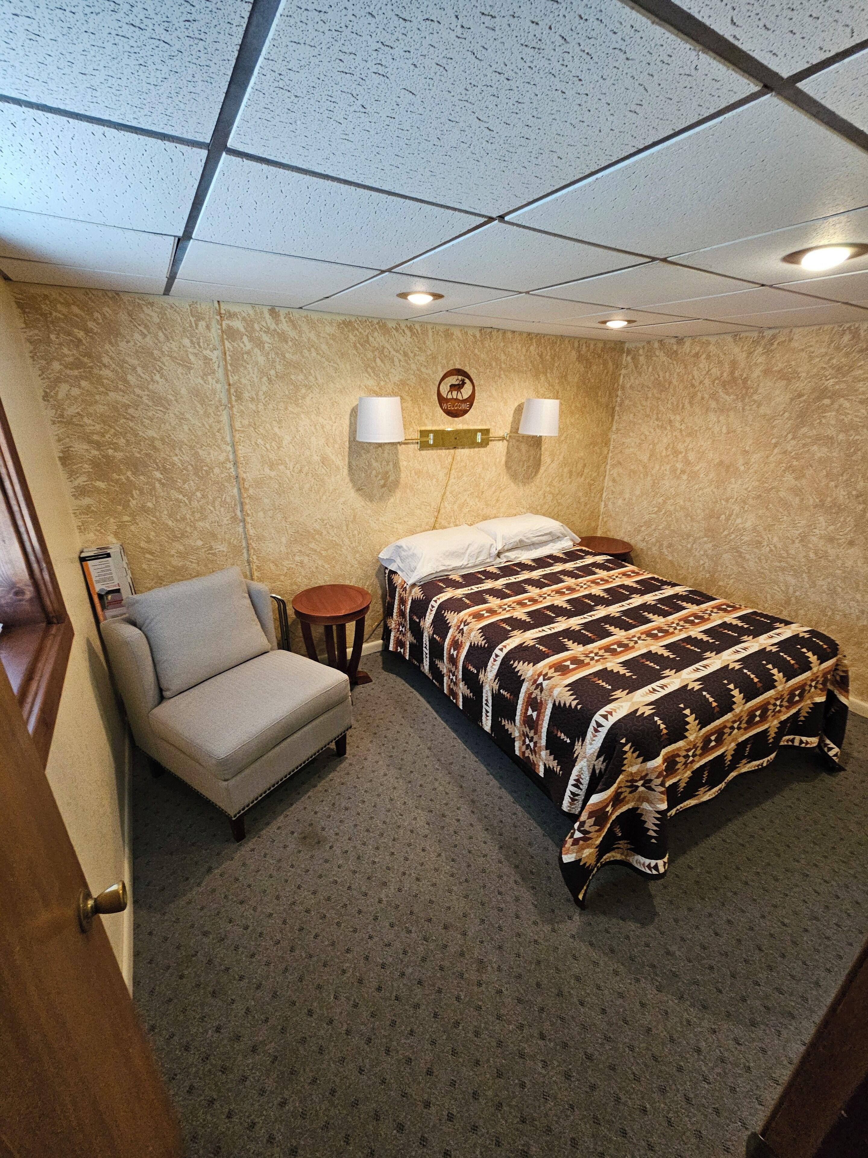 A hotel room with a bed, a chair, a small table, and a wall-mounted lamp.