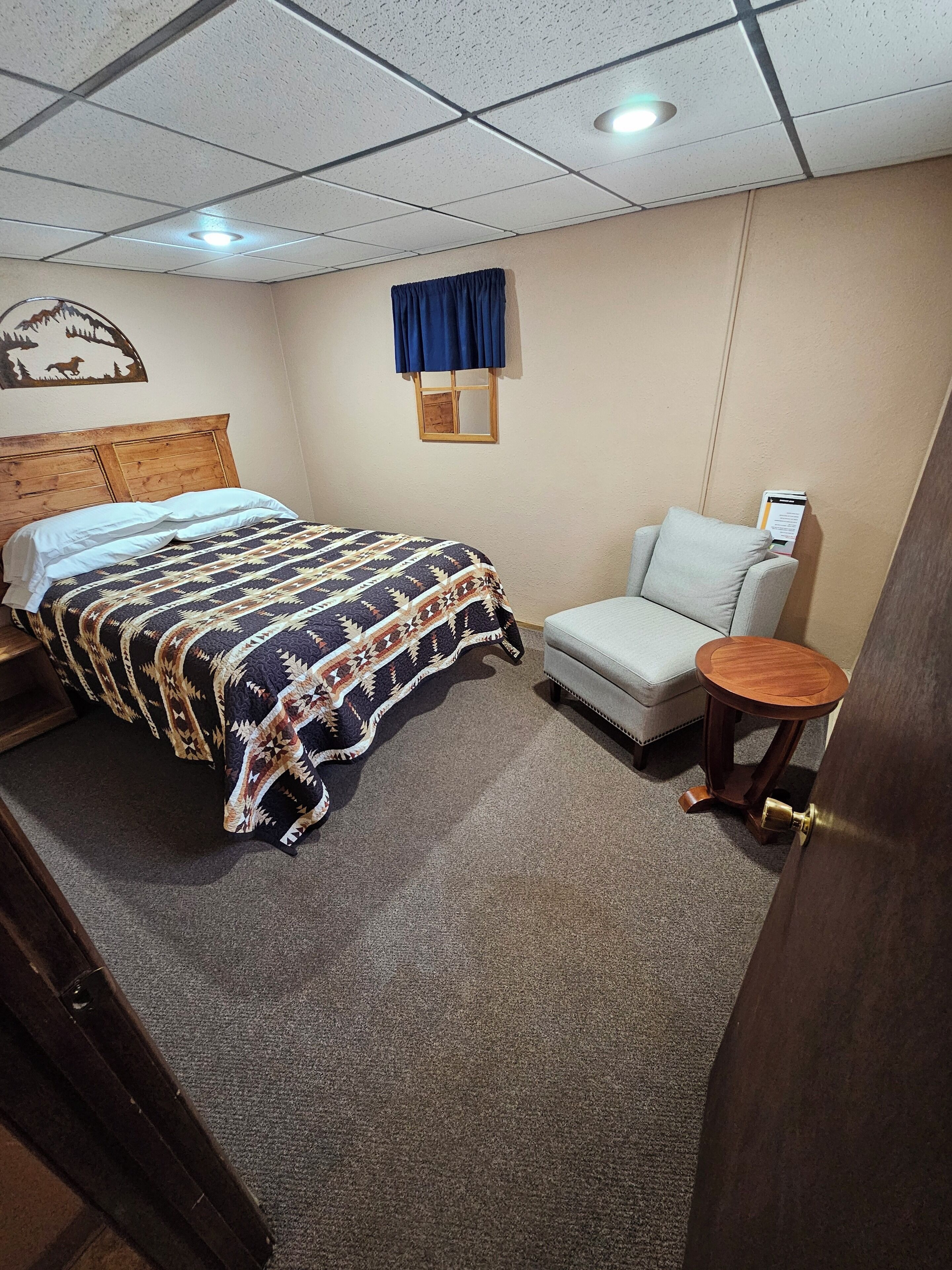 Habitación de hotel con cama, una silla, una mesita, ventana con cortinas y un adorno en la pared.