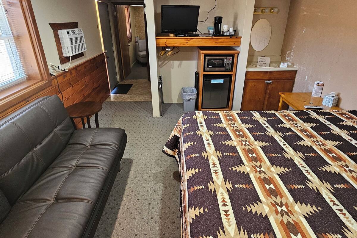 A compact hotel room with a bed, a sofa, a kitchenette, and a television.