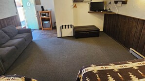 Una habitación con sofá, cama con ropa de cama estampada, una mesita con una silla, microondas y televisor montado en la pared.