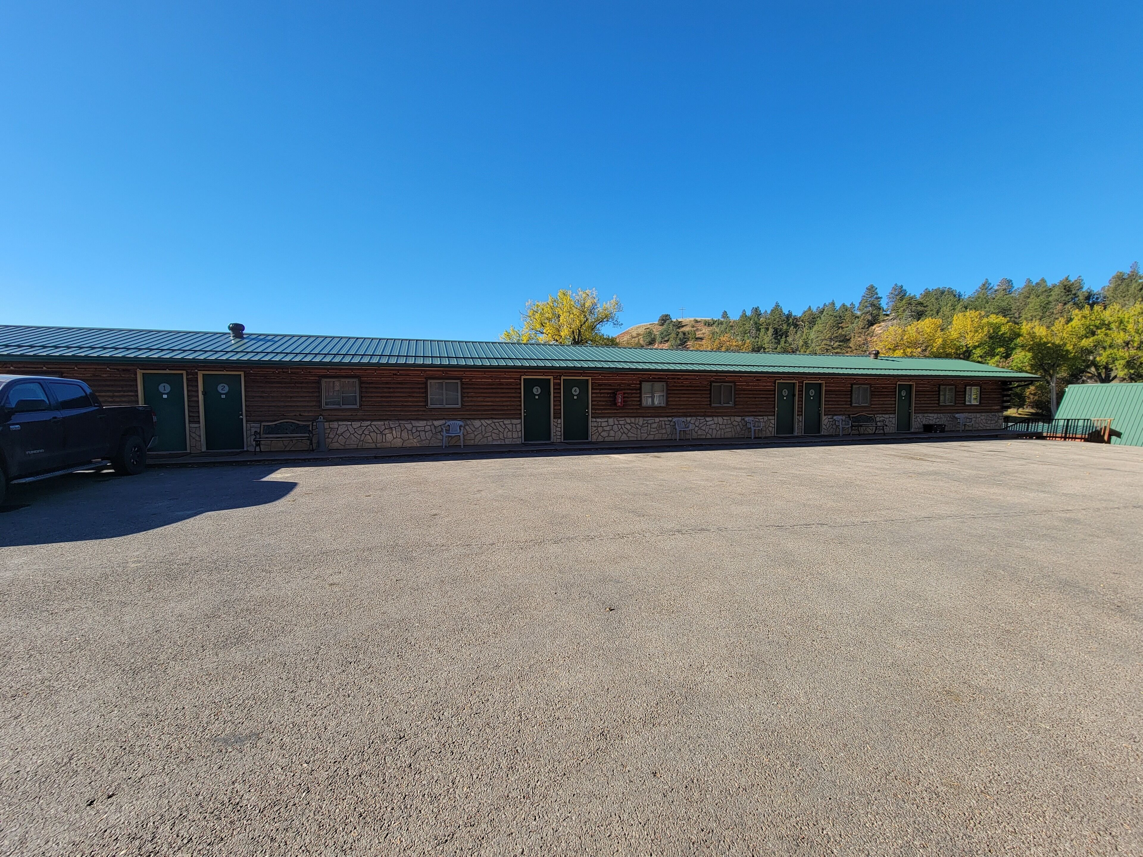 A building with a green roof, wooden exterior, and stone accents. It has multiple doors and windows, and a gravel parking area in front.