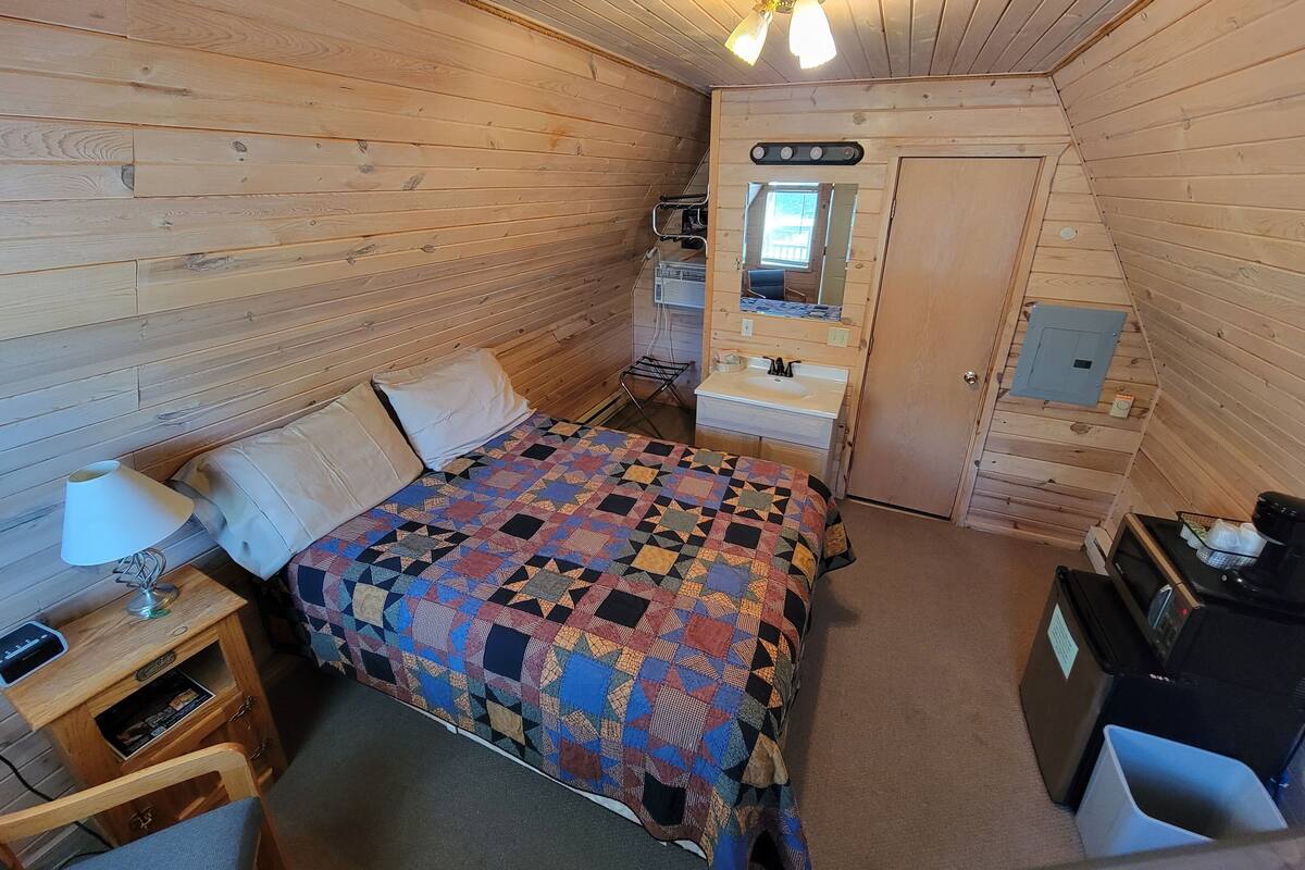 A compact bedroom with a wooden floor, a bed with a colorful quilt, a bedside table with a lamp, a small desk, and a mirror above the sink.