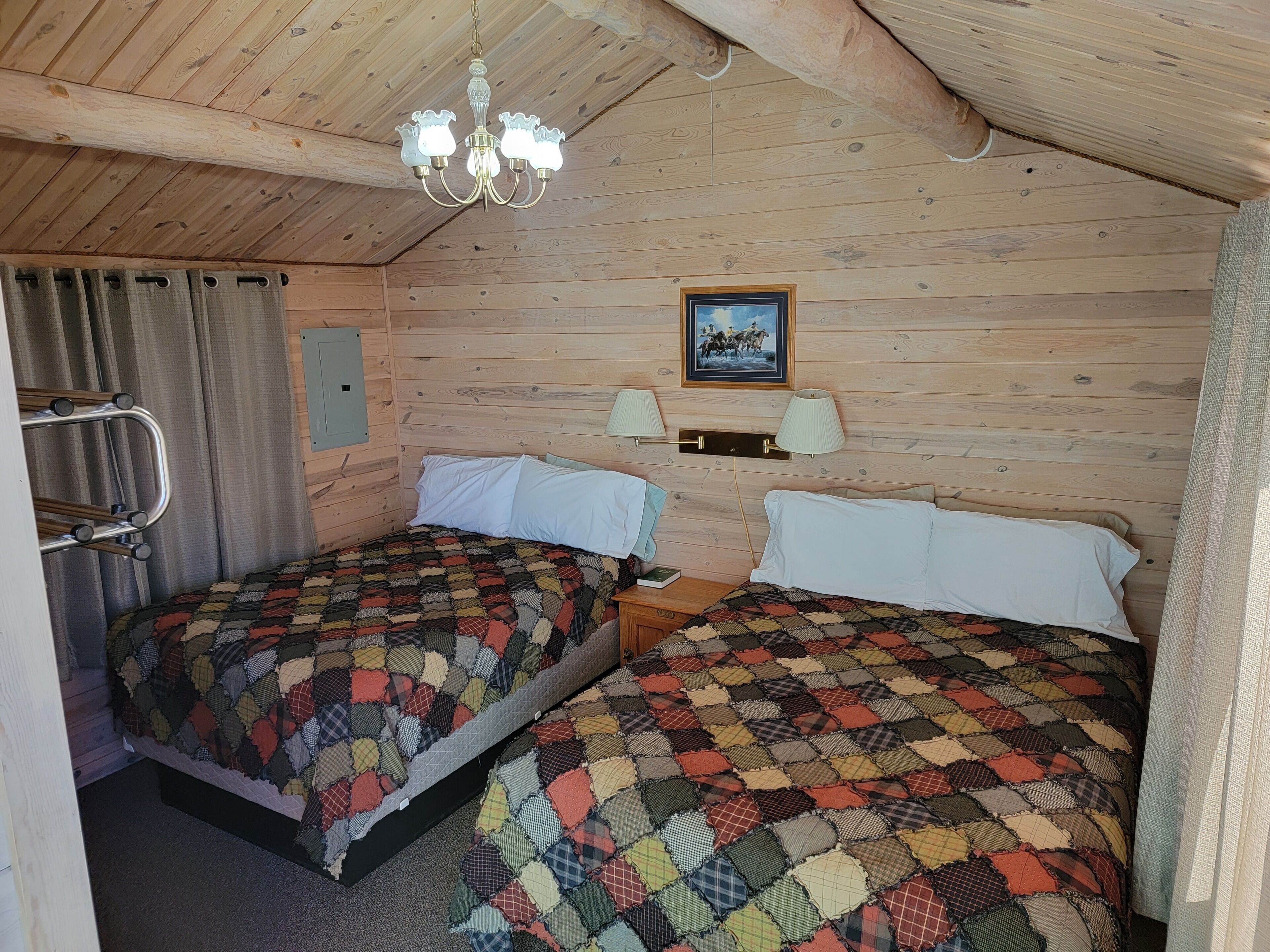 A room with two beds, wooden walls, a chandelier, a framed picture, and a nightstand with lamps.