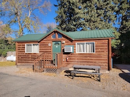 A wooden cabin with a green roof, a front door, and a small porch with a bench.