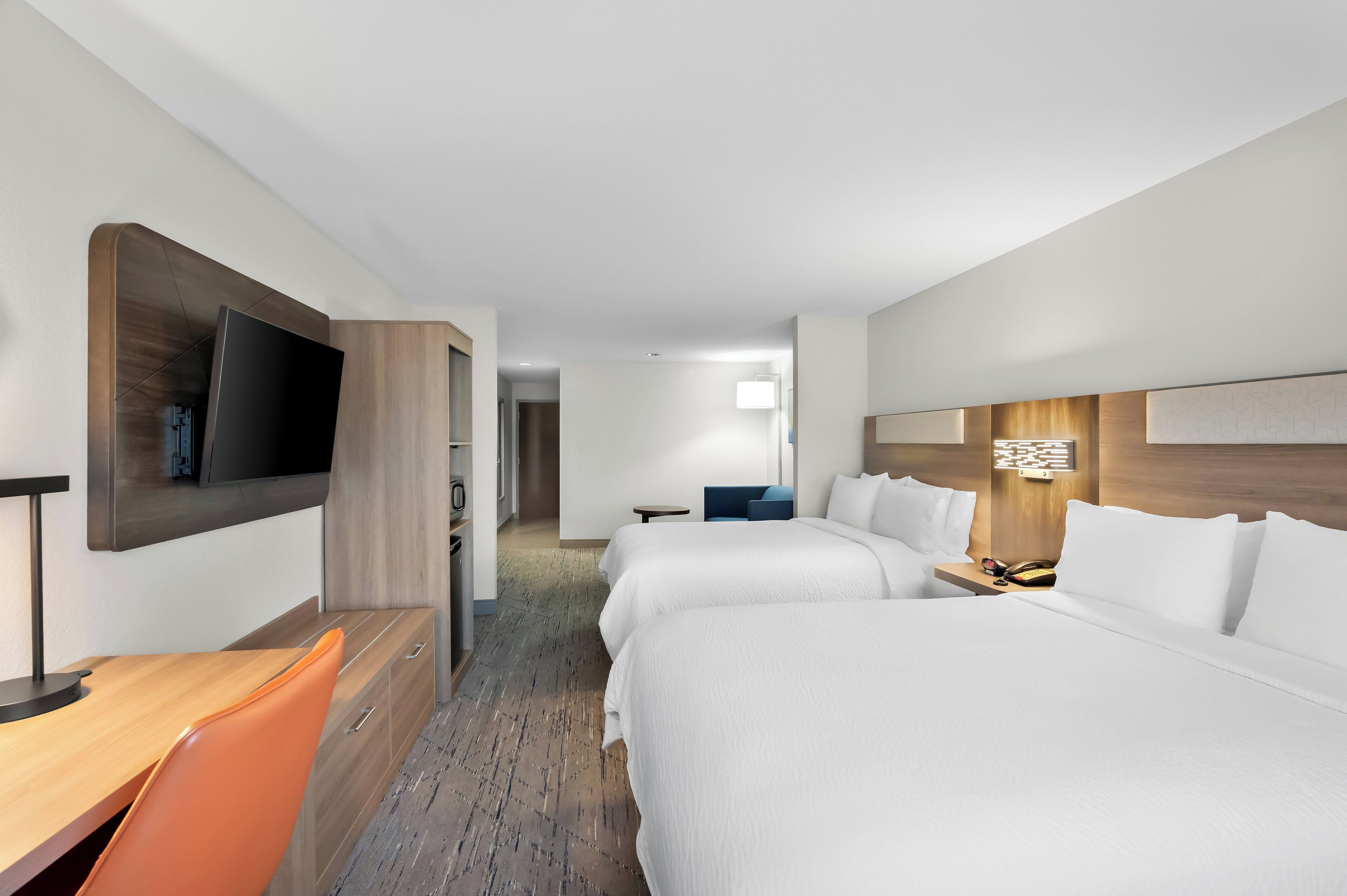 A hotel room with a flat-screen TV, two beds, a desk with a chair, and a small table with a lamp.
