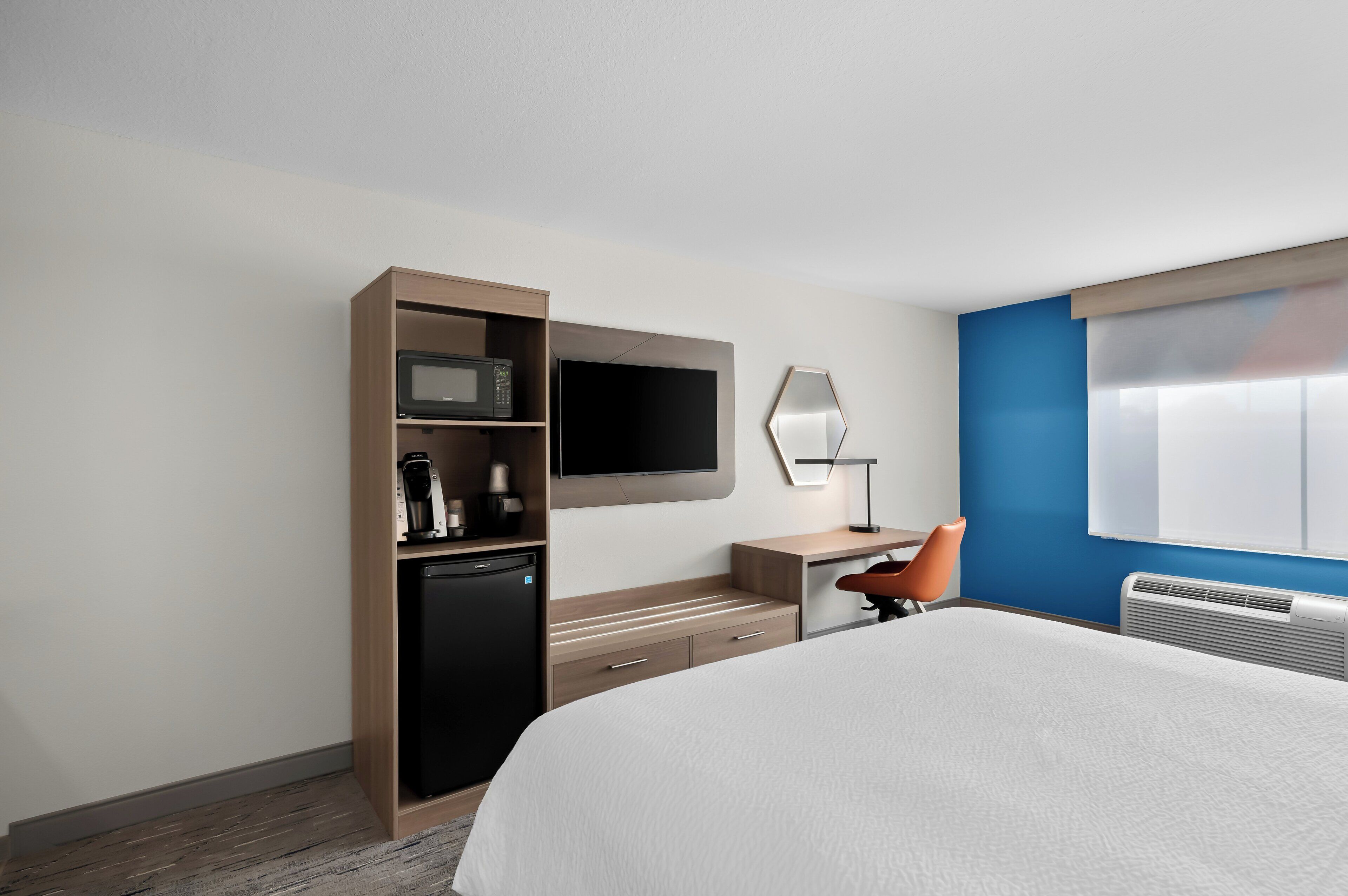 A hotel room with a bed, a desk with a chair, a microwave, a coffee maker, a TV, and a heater.