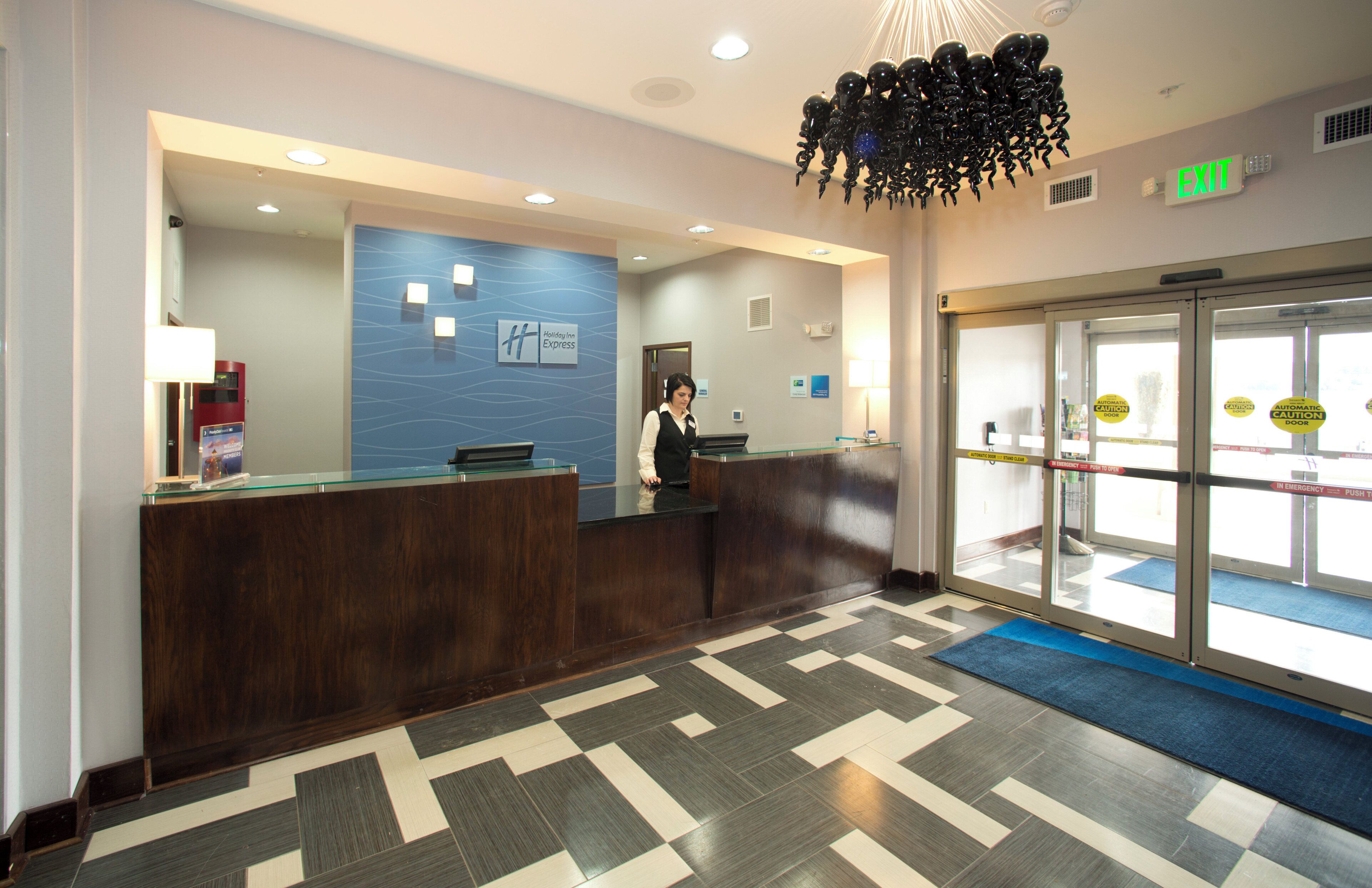 Área de recepção de hotel com recepcionista, placa Holiday Inn Express, sinal de saída e balcão de check-in.