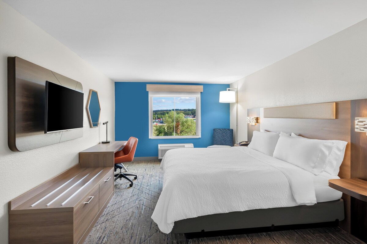 A modern hotel room with a large bed, a desk, a flat-screen TV, and a window with a city view.