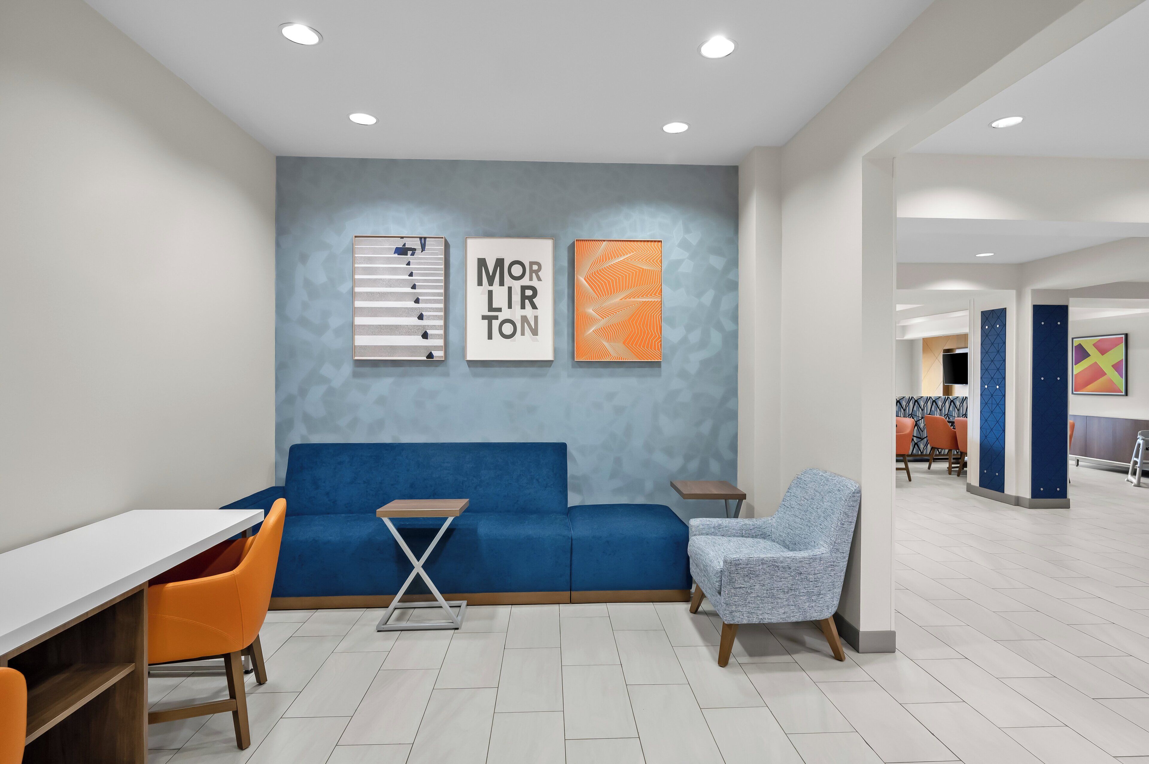 A modern lounge area with a blue sofa, orange chairs, a small table, and artwork on the wall.