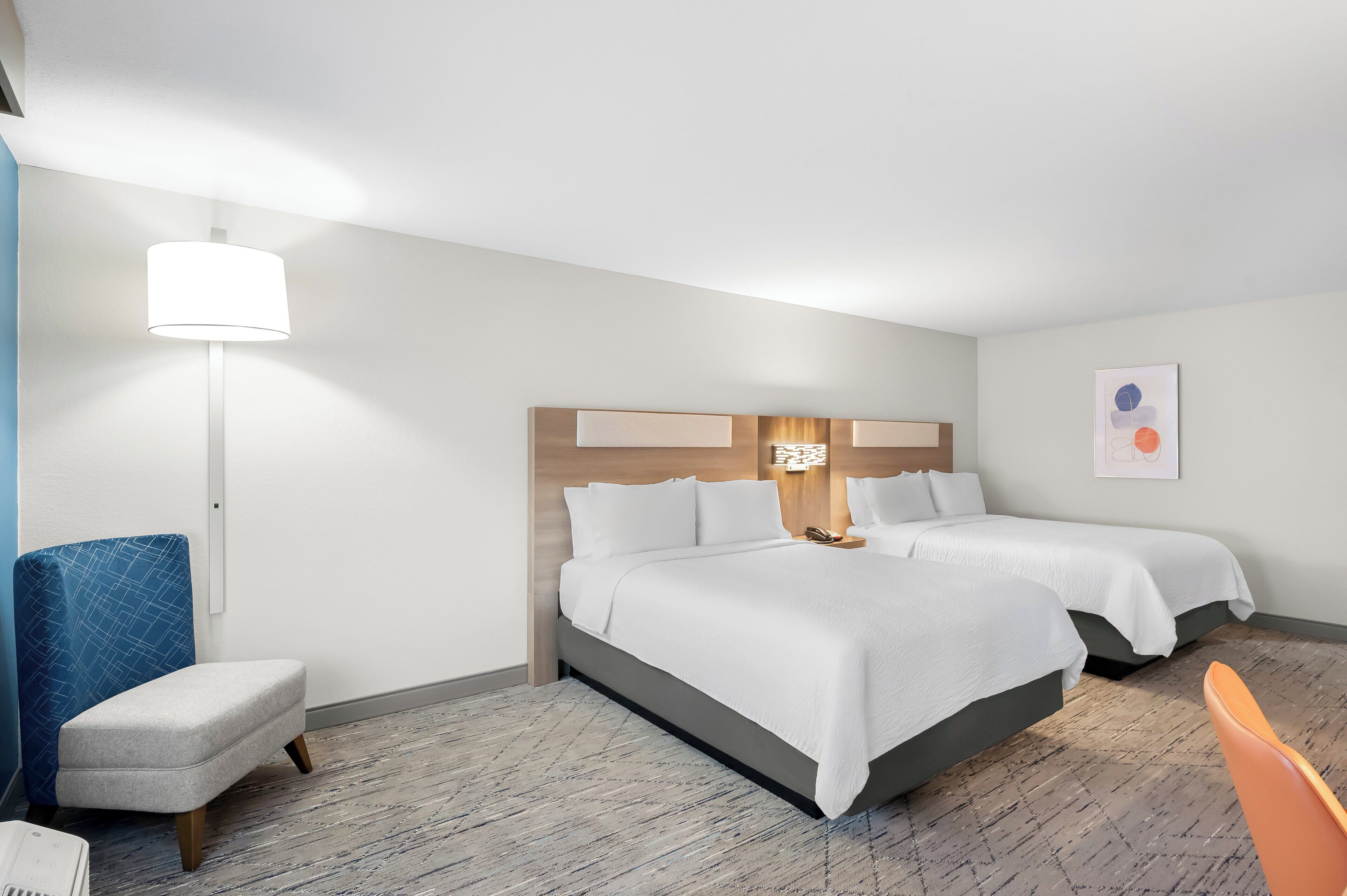A hotel room with two beds, a desk, a chair, a lamp, and a wall art piece.