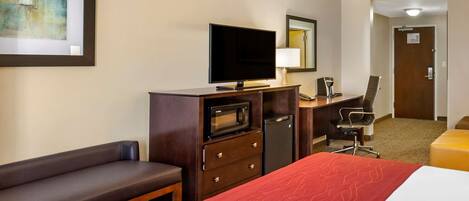 A hotel room with a bed, a desk with a computer, a television, a microwave, a refrigerator, a chair, and a bench.
