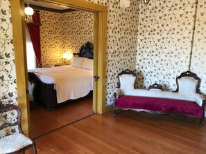 Una habitación con piso de madera, una cama con ropa de cama blanca, una cortina roja, una mesita de noche, una lámpara, una silla y un sofá con una manta roja.