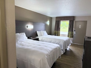 Habitación de hotel con dos camas, una mesita de noche, un escritorio, una silla, una ventana con cortinas y una puerta.