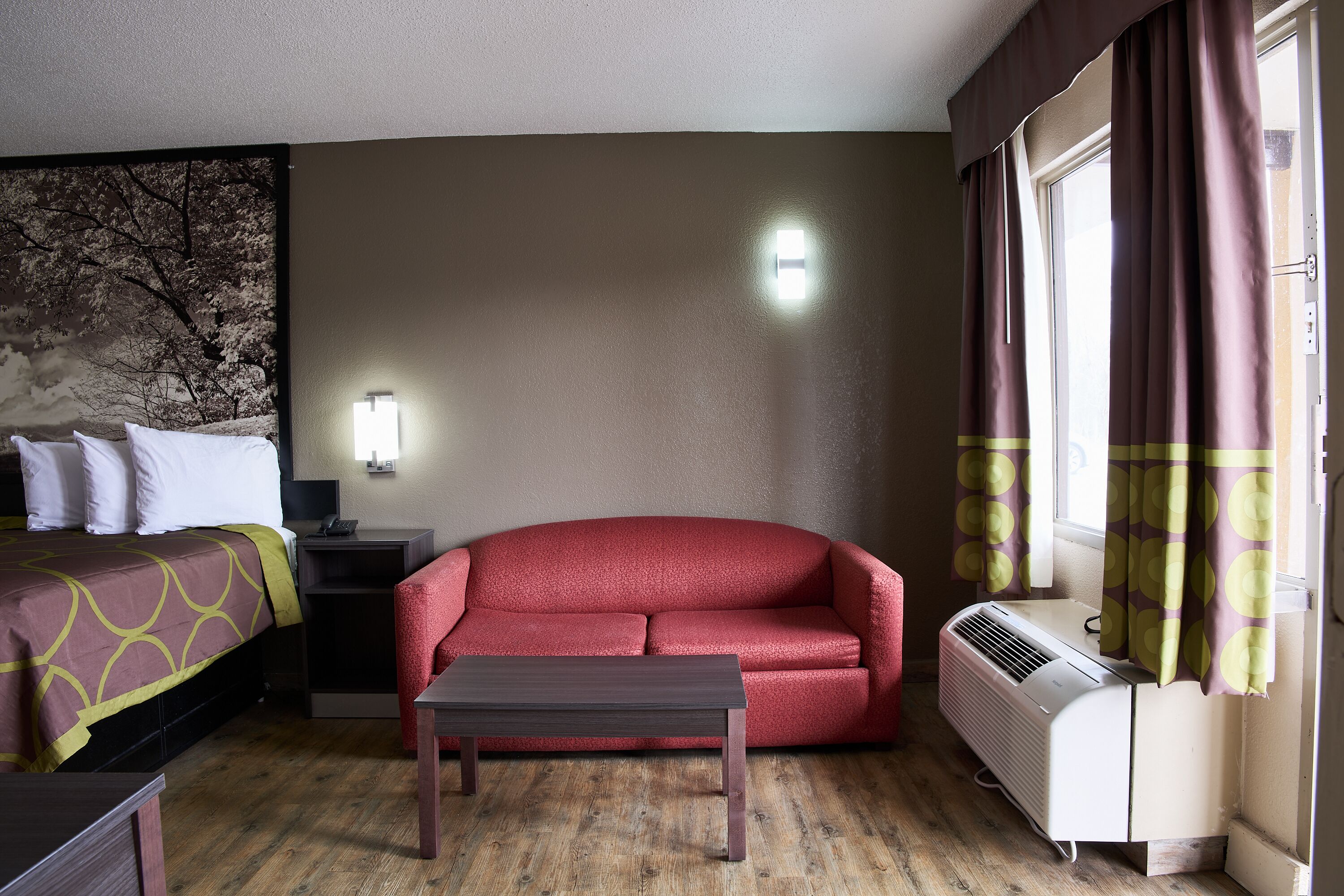 A hotel room with a bed, a red sofa, a small table, a heater, and a window with curtains.