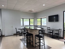 A spacious room with a central island table surrounded by chairs, large windows with a view of the outdoors, and a flat-screen TV mounted on the wall.