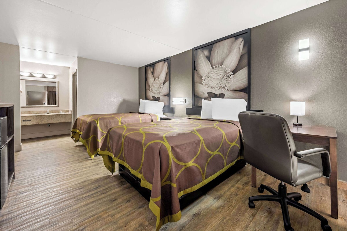 A hotel room with two beds, a desk, a chair, a mirror, and a large floral artwork on the wall.