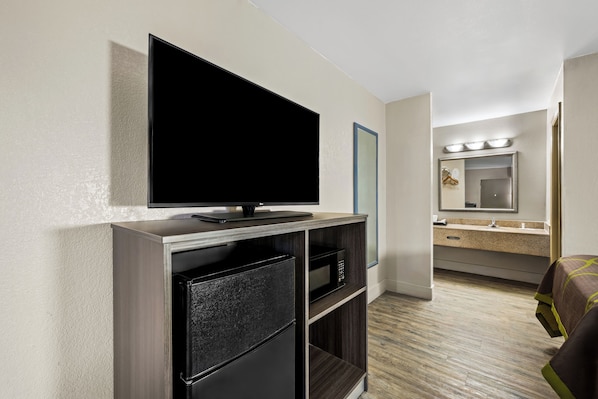 A hotel room with a flat-screen TV on a wooden stand, a microwave, a small refrigerator, a bathroom with a sink and mirror, and a bed with a brown blanket. - Quality Inn (Garysburg)