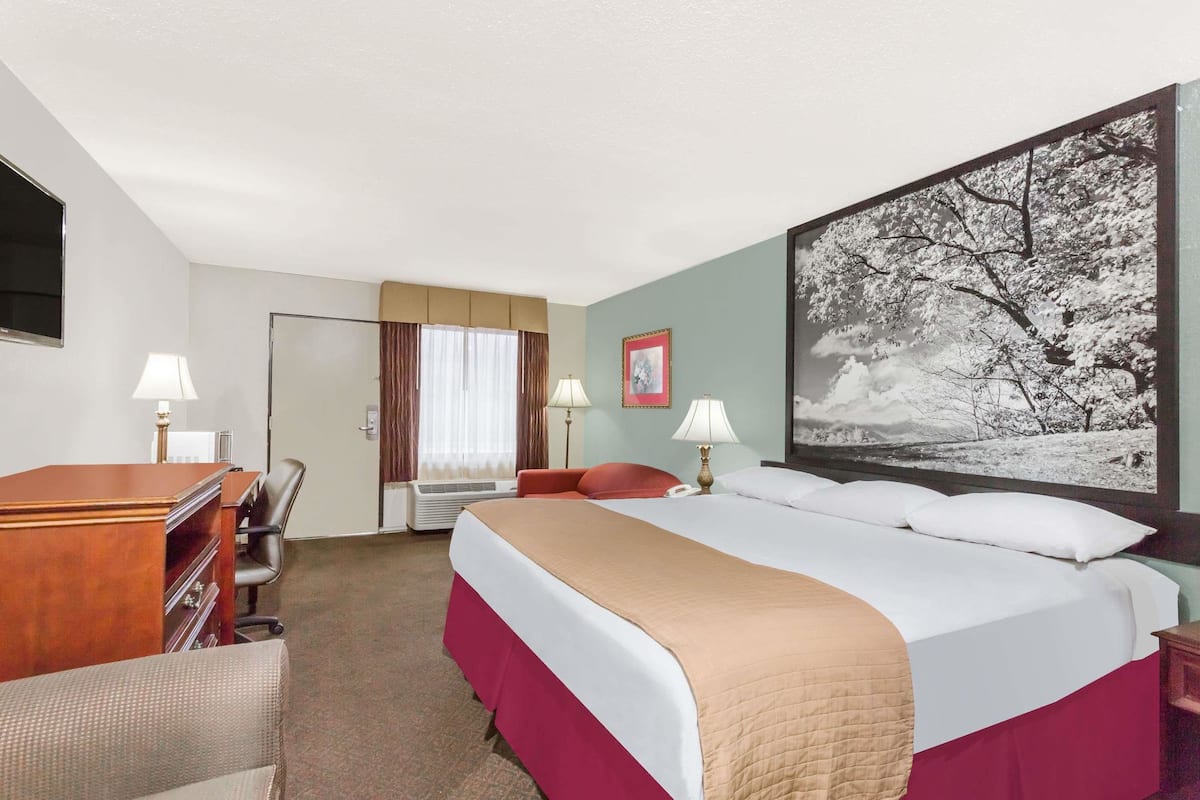 A hotel room with a bed, desk, chair, lamps, and a large framed picture.