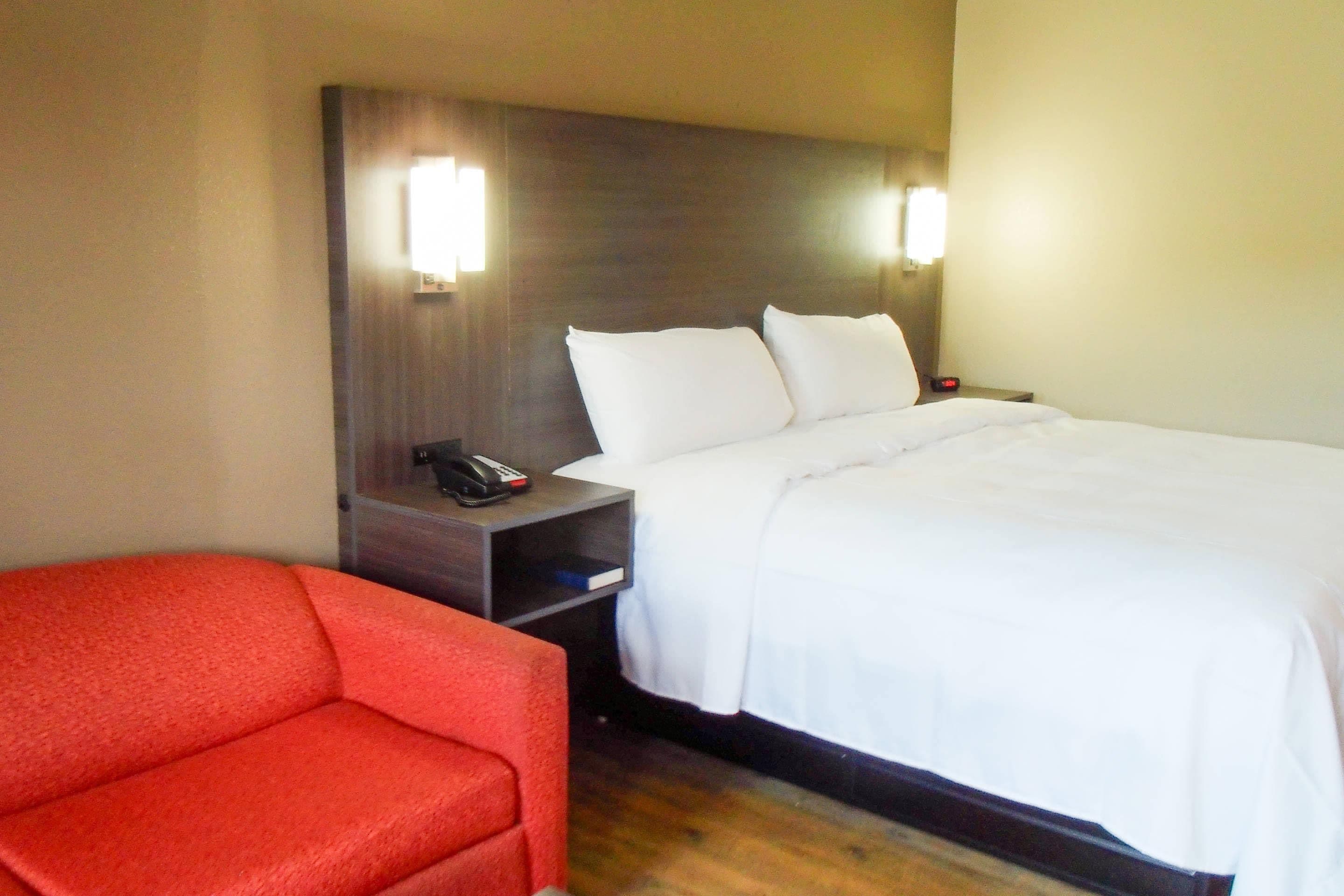 A hotel room with a bed, a nightstand, a telephone, and a red chair.