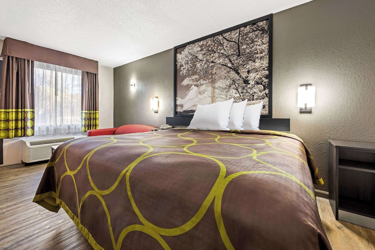 A hotel room with a large bed, a window with curtains, a wall-mounted artwork, and a bedside table.