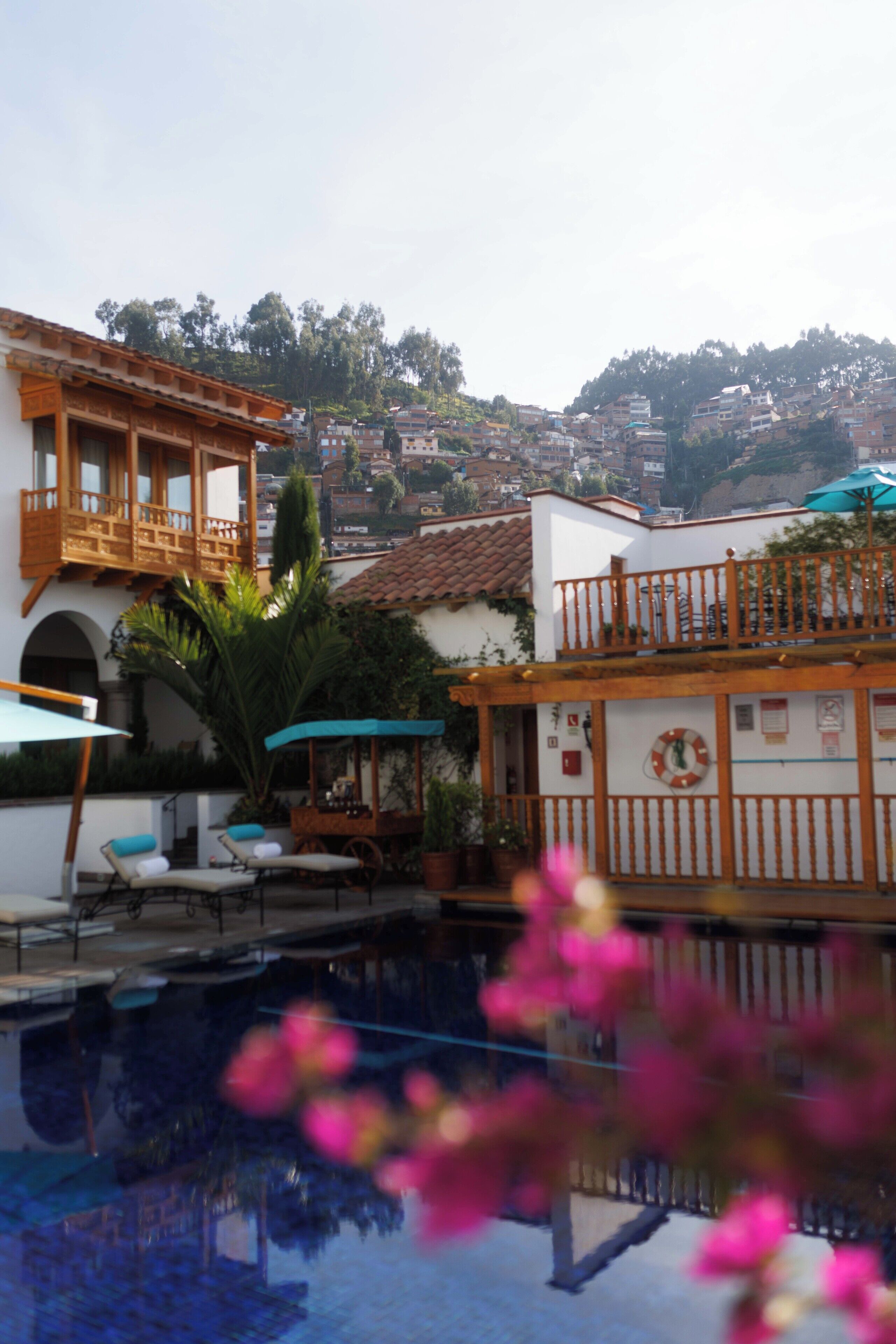 A pool area with lounge chairs and umbrellas, a building with a balcony, and a view of a hillside with houses.