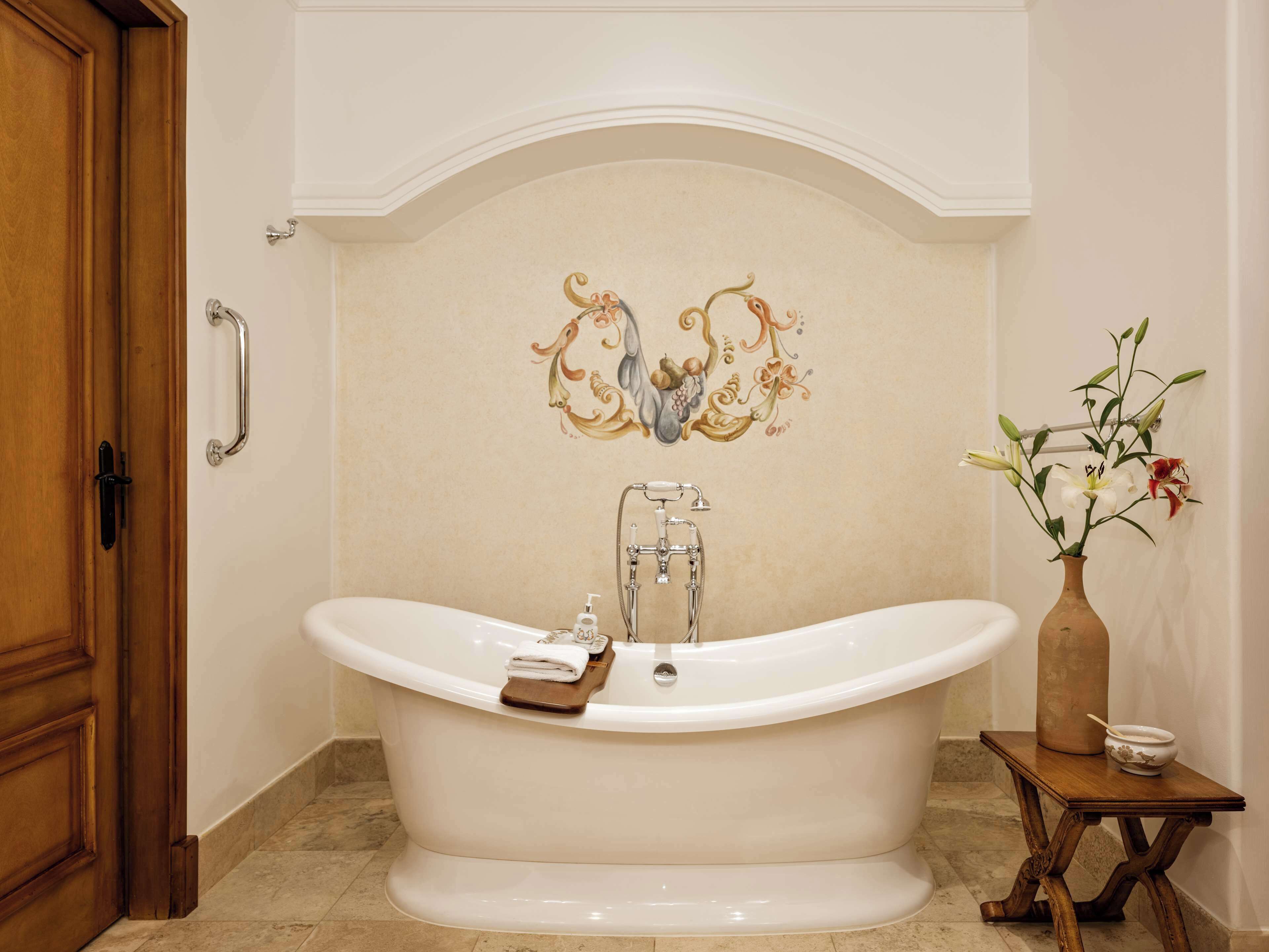 Una bañera independiente con una toalla y dispensador de jabón, una puerta de madera y un florero con flores sobre una mesita.