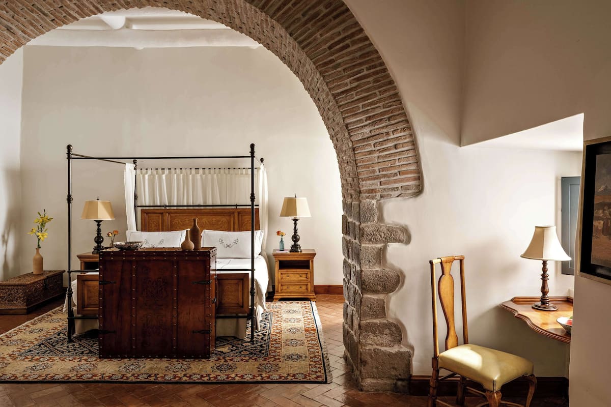 A bedroom with a four-poster bed, a wooden chest of drawers, a desk with a chair, and a stone archway.