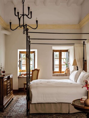 Un dormitorio con una cama grande, un aparador de madera, un escritorio con una silla, dos ventanas con cortinas y una araña.
