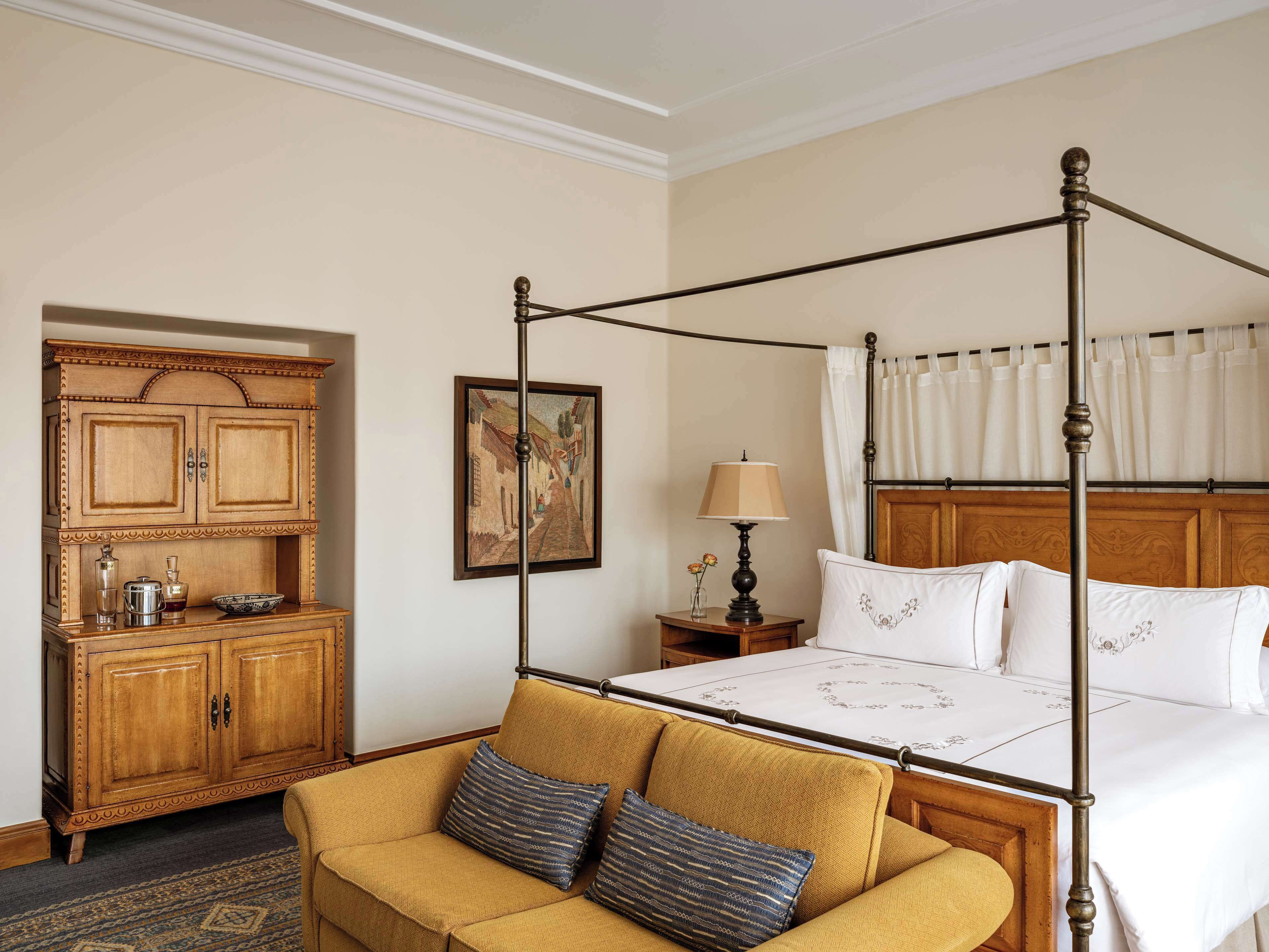 A four-poster bed with a wooden headboard, a sofa with patterned cushions, a wooden cabinet with decorative trim, and a framed painting on the wall.