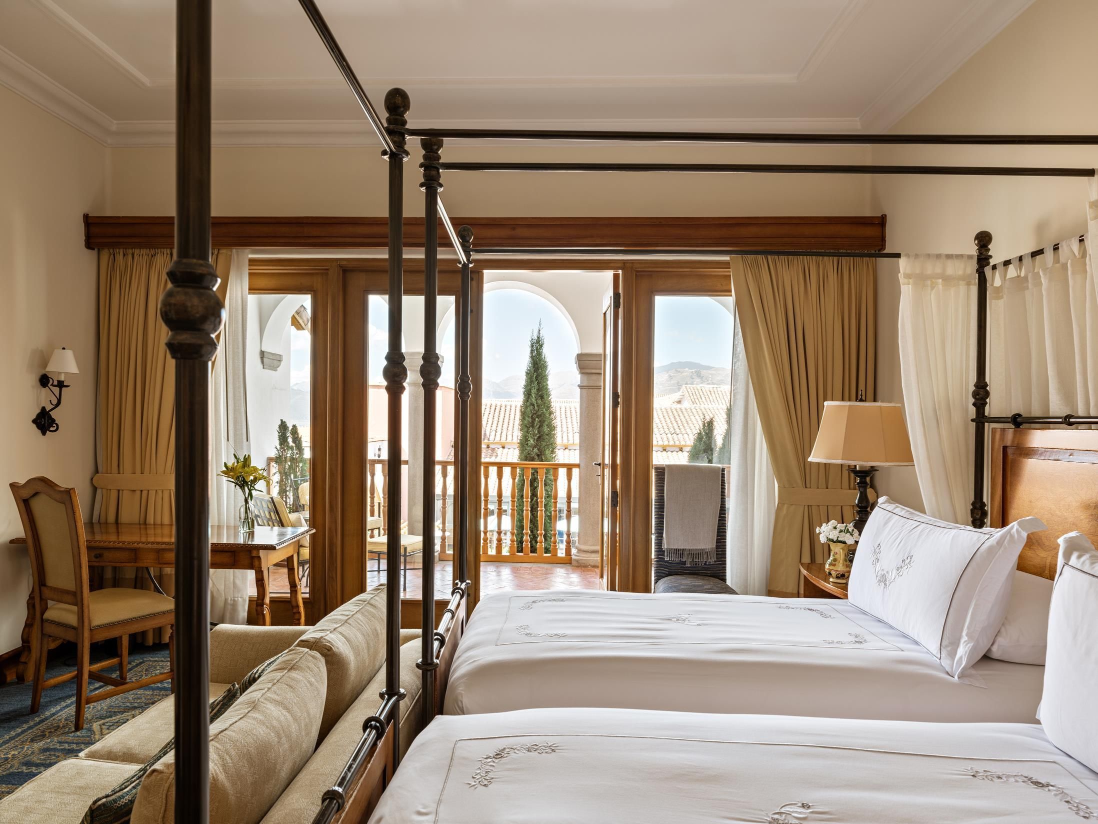 A four-poster bed with a balcony view, a wooden desk with a chair, and a window with curtains.