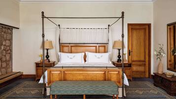 A four-poster bed with a wooden headboard, two bedside tables with lamps, a bench at the foot of the bed, a stone wall, and a wooden door.