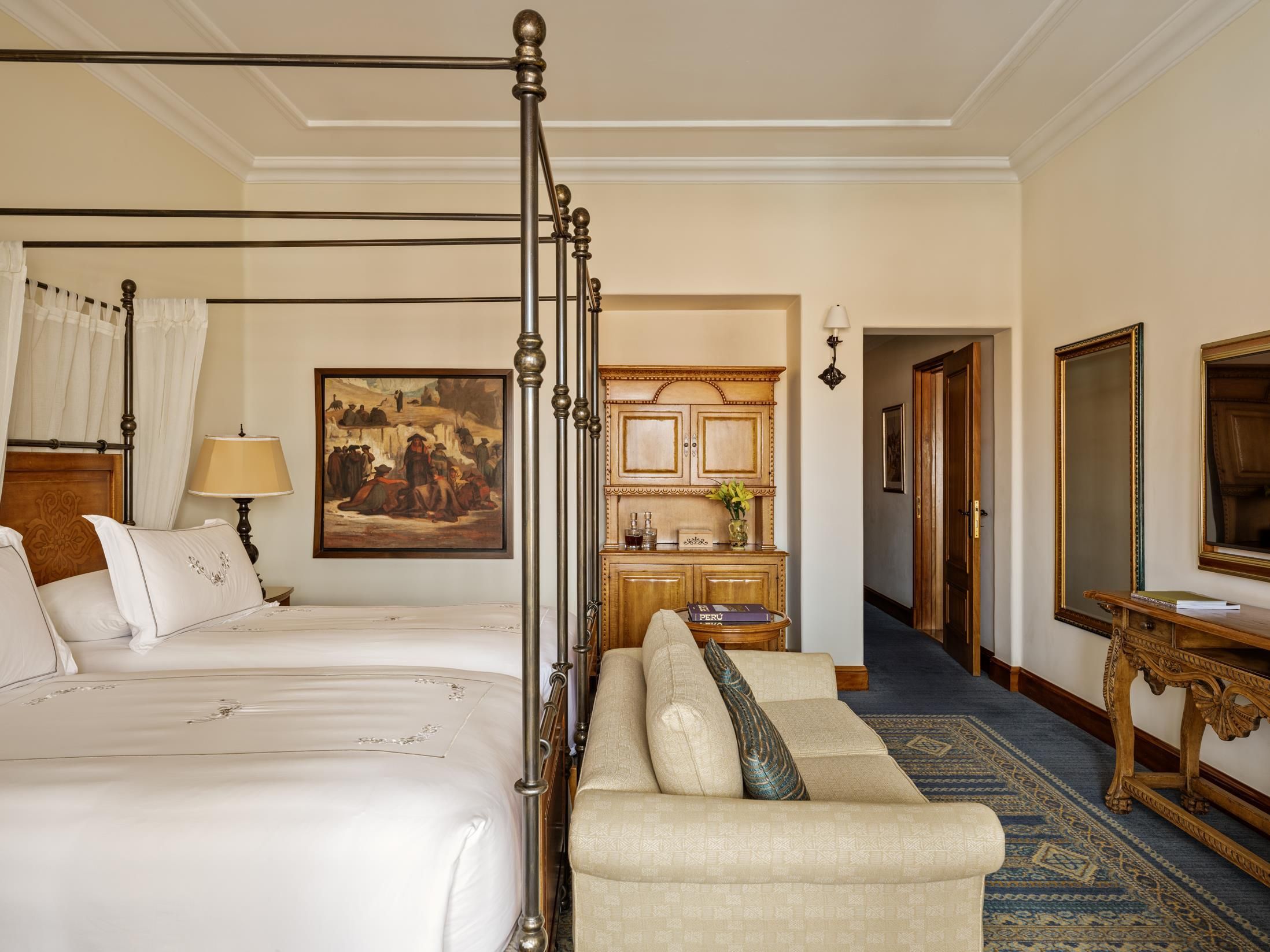A hotel room with a four-poster bed, a sofa, a painting, a wooden cabinet, and a mirror.