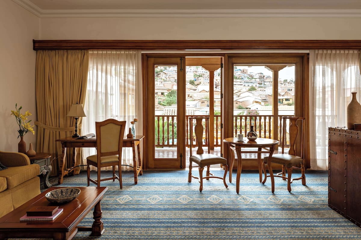 A room with a blue patterned carpet, wooden furniture, a balcony with a view of a hillside, and a vase with flowers on a side table.