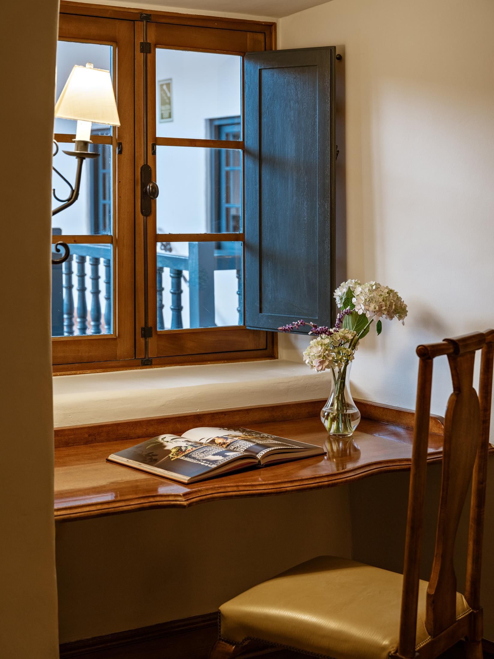 A wooden desk with a chair, a window with a view, and a vase with flowers.