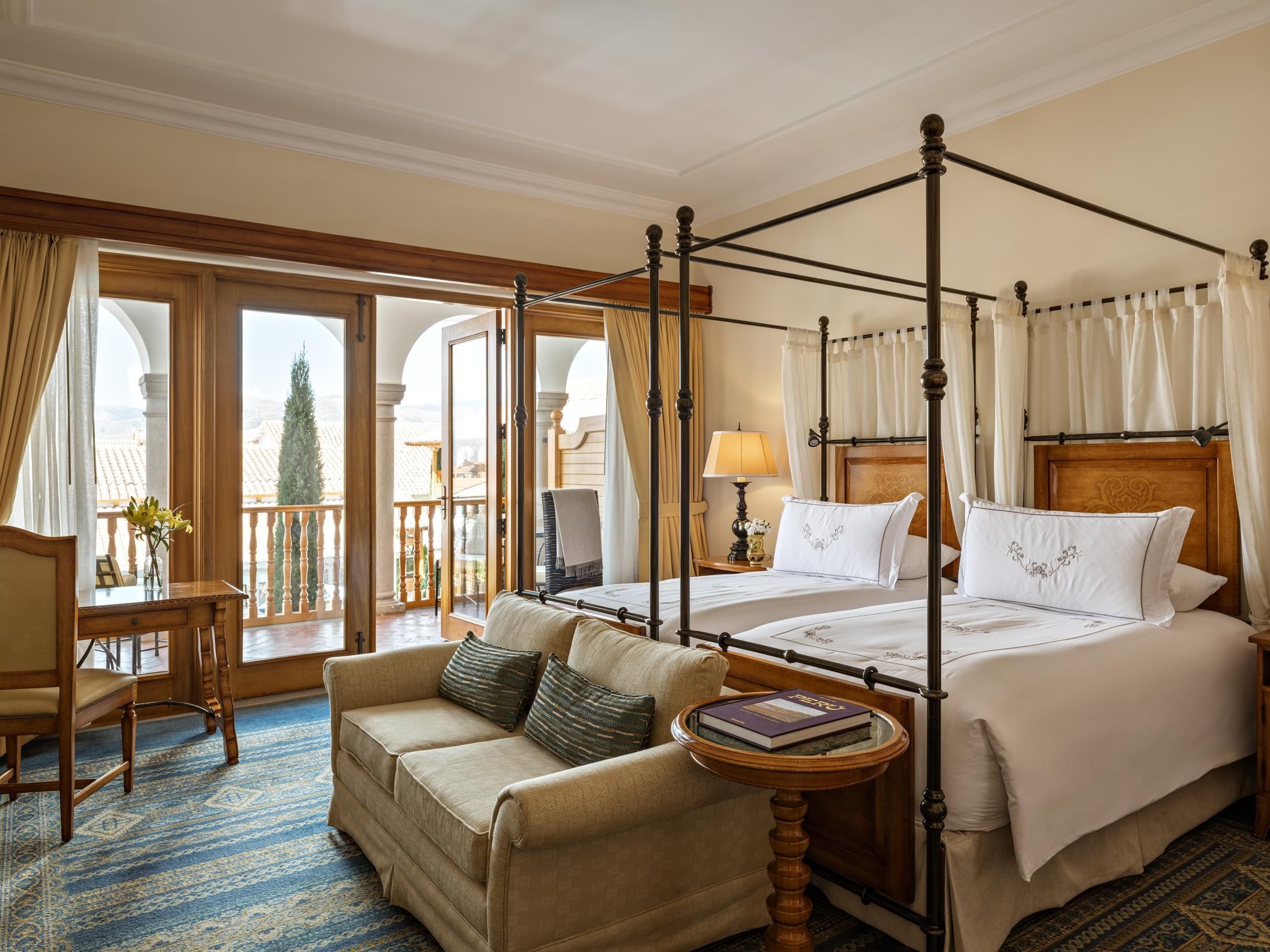 A four-poster bed with a wooden headboard, a sofa, a small round table with a book, and a view of the outdoors through a glass door.