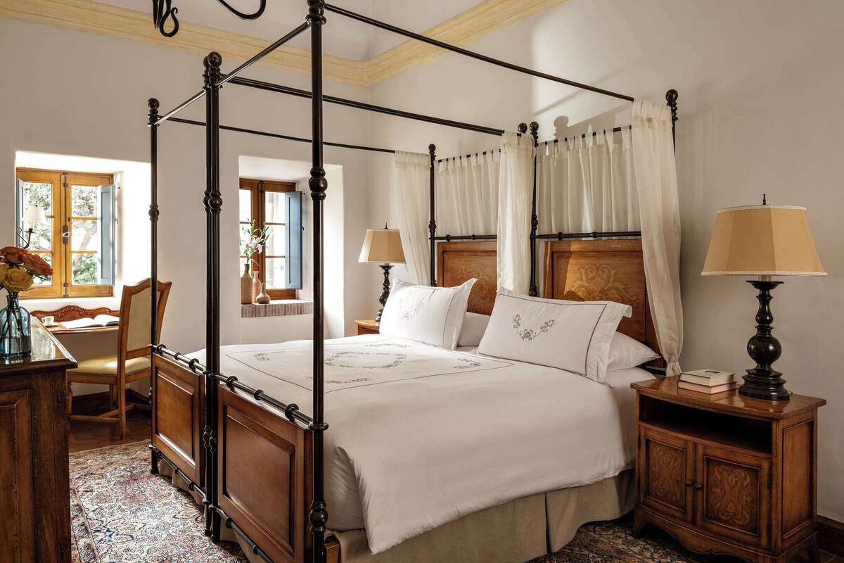 A four-poster bed with a wooden headboard, a bedside table with a lamp, and a chair with a vase of flowers.