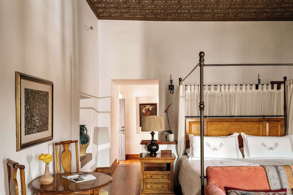 A bedroom with a four-poster bed, a wooden nightstand, a table with chairs, and a chair with a cushion.