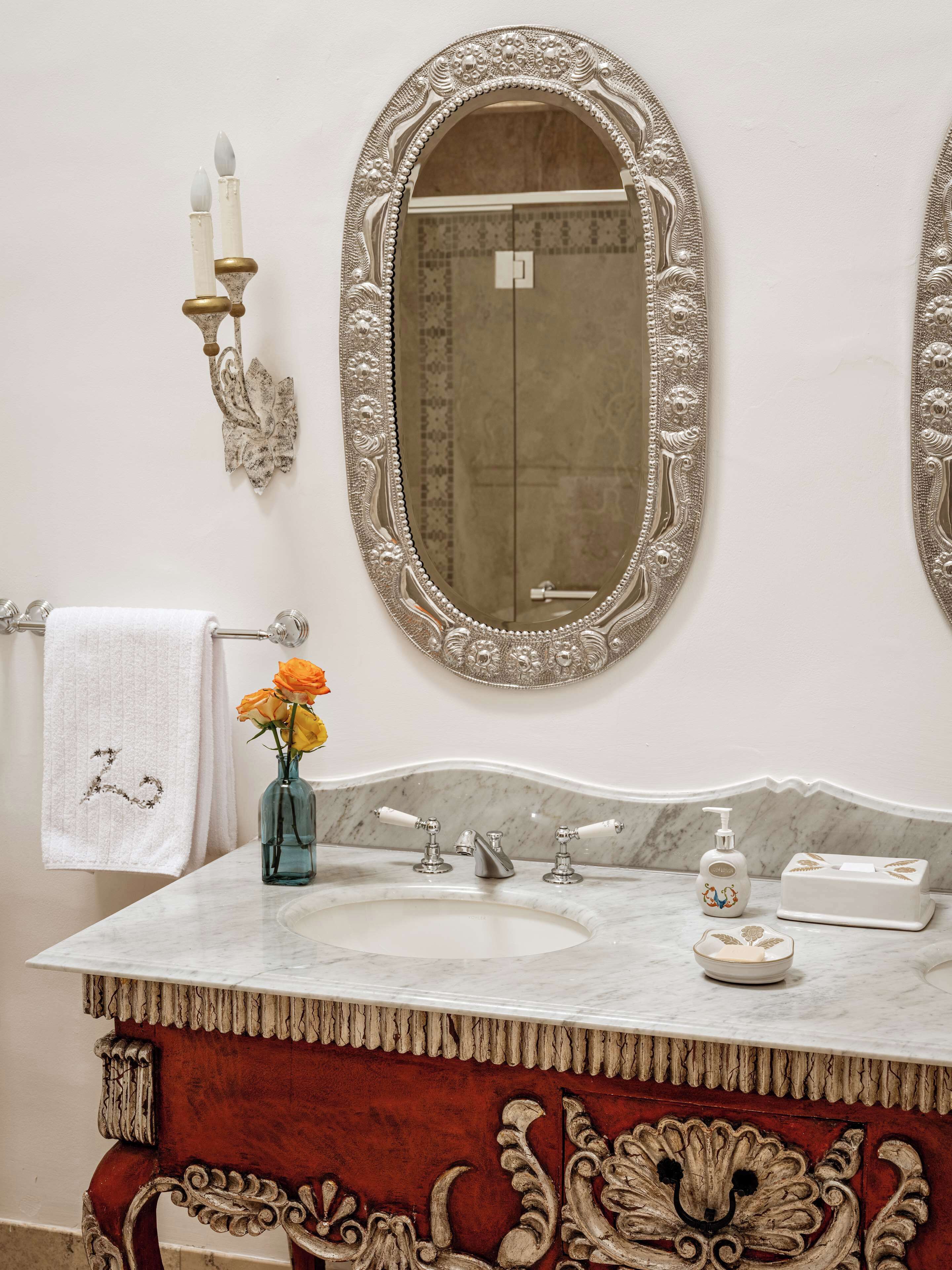 A bathroom with an ornate mirror, a marble sink, a red vanity, and a towel with a monogram.
