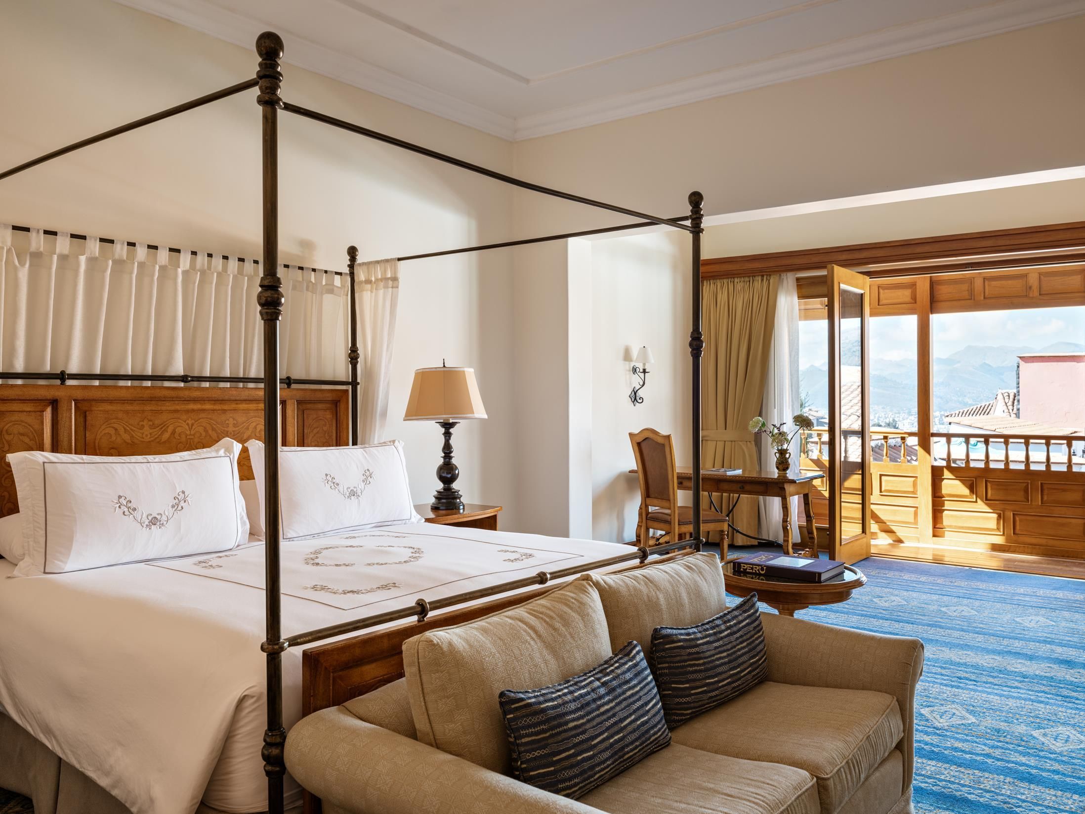 A four-poster bed with a wooden headboard, a bedside table with a lamp, a sofa with cushions, and a view of a balcony with a table and chairs.