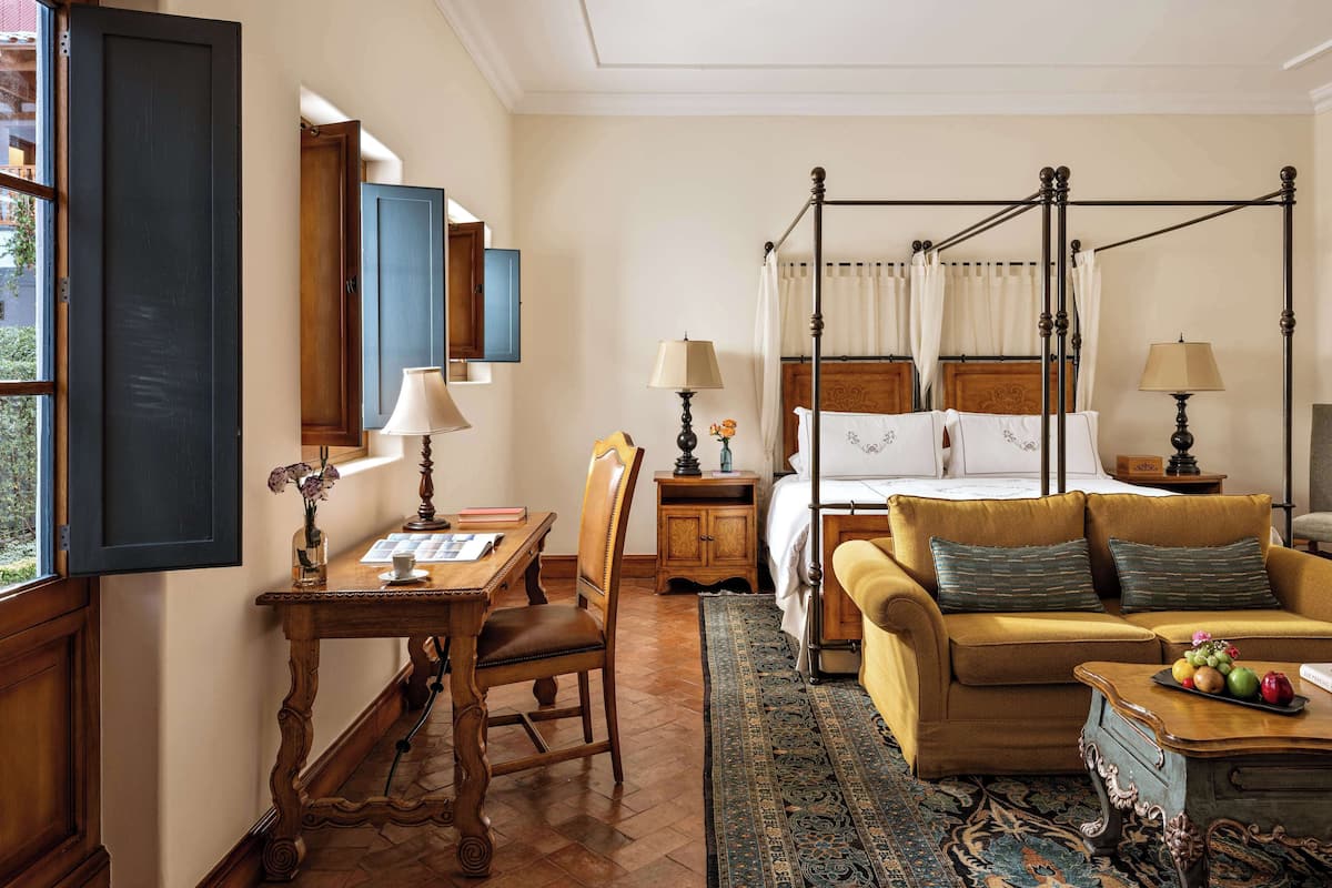 A bedroom with a four-poster bed, a wooden desk with a lamp, a chair, a sofa, and a coffee table with fruit.