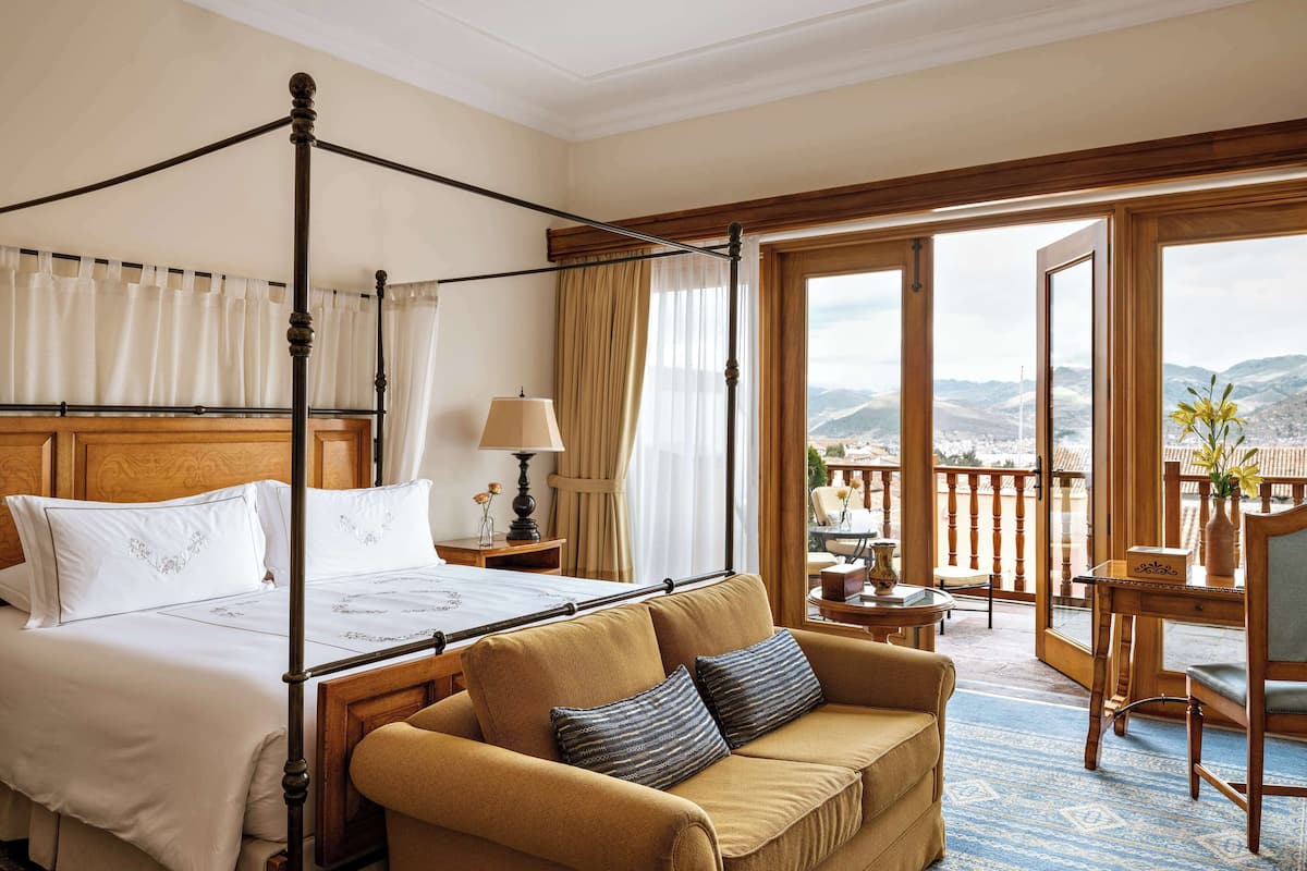 A hotel room with a four-poster bed, a sofa, a small table with a lamp, and a view of the mountains through large windows.
