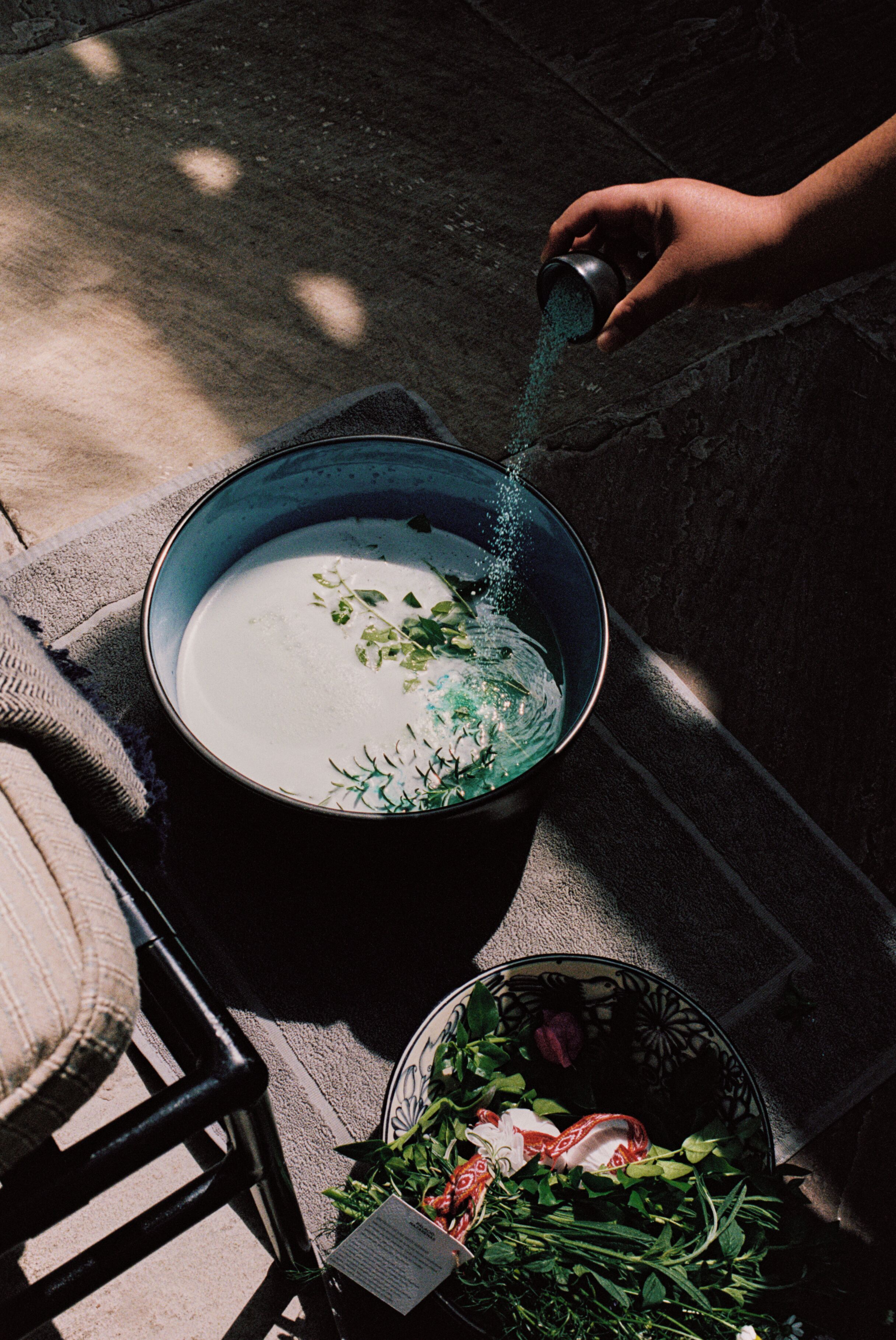A large pot on a table with a person pouring a liquid into it. There is a bowl with greens and a tag on the table.