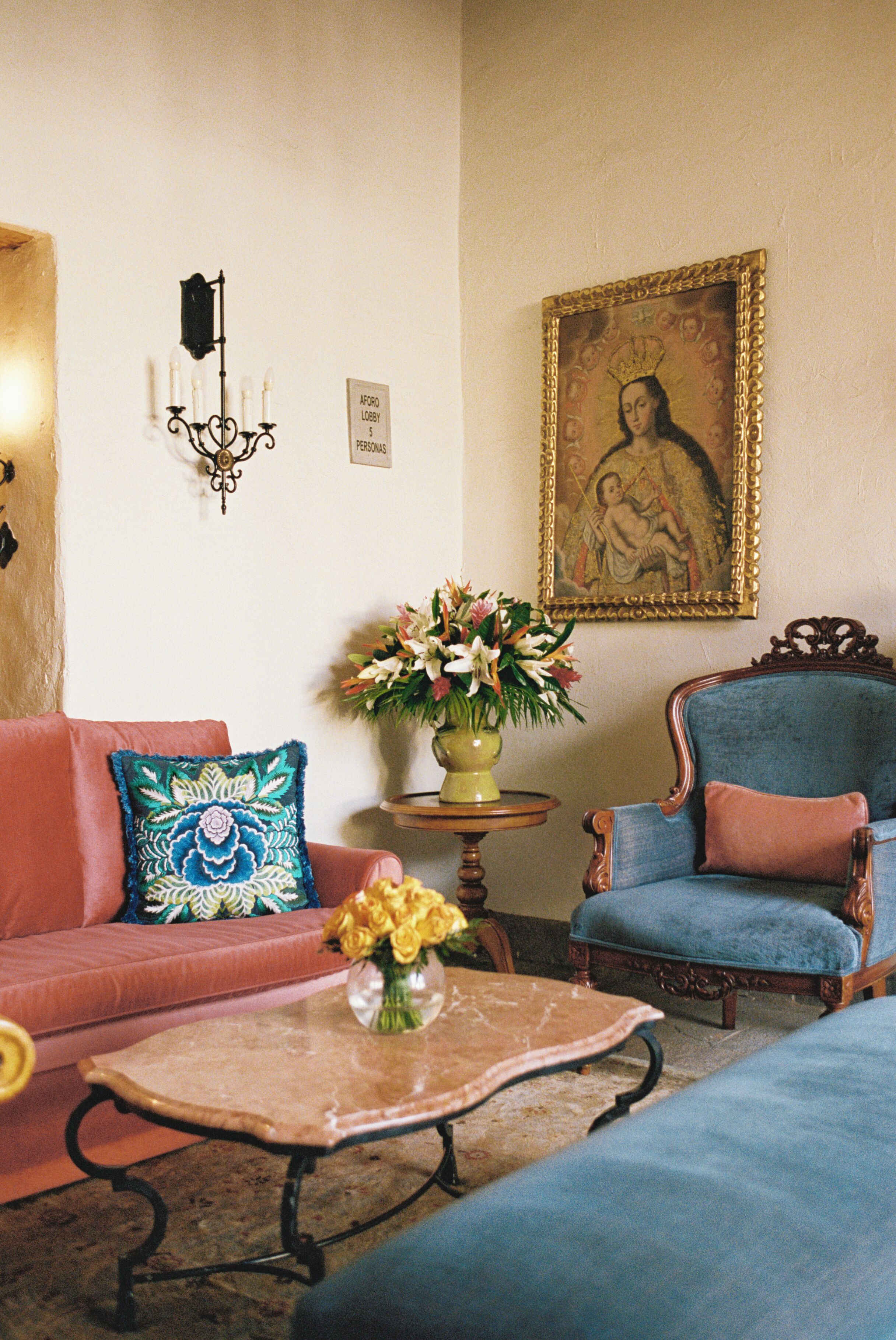 A room with a red sofa, a blue armchair, a coffee table with a vase of flowers, a wall-mounted lamp, and a framed painting.