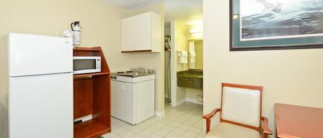 A compact kitchenette with a refrigerator, microwave, and sink. There is a small dining area with a chair and table.